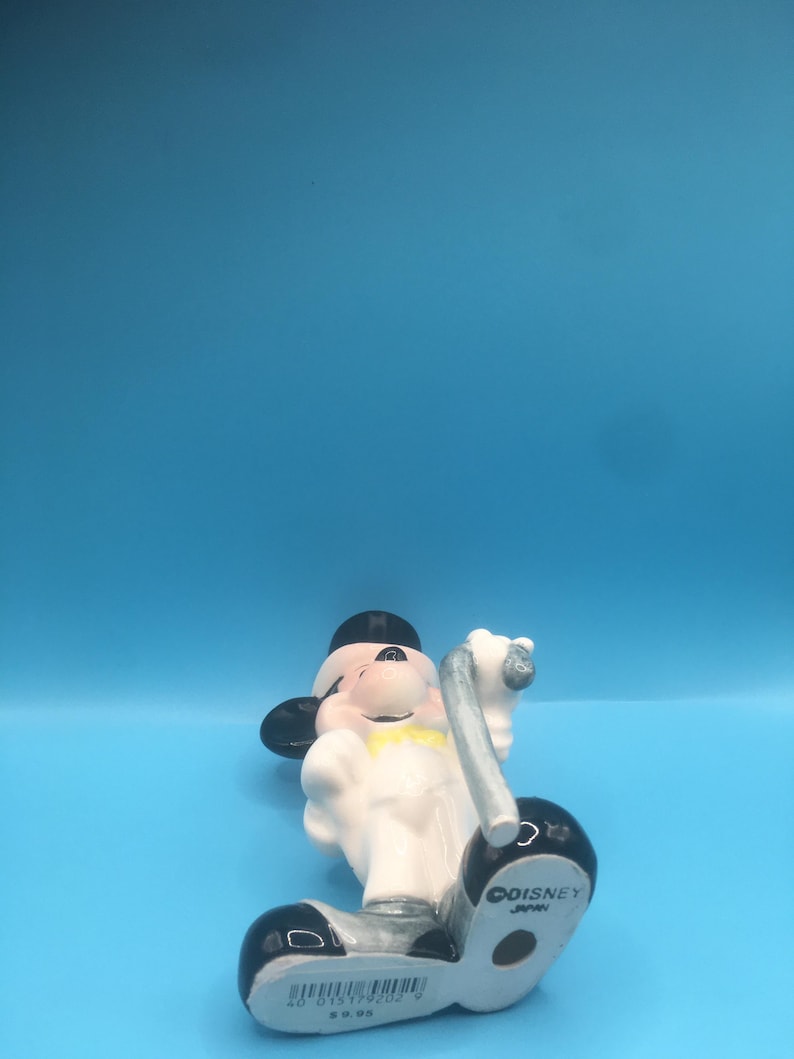 May include: A white ceramic figurine of Mickey Mouse dressed as a conductor, holding a baton. He is wearing a black top hat, a white shirt with a yellow bow tie, and black shoes. The figurine is made in Japan and has the Disney logo on the bottom.
