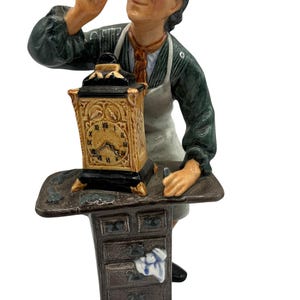 May include: A figurine of a clockmaker examining a small clock. The clock is gold and black with Roman numerals. The clockmaker wears a green shirt, white apron, and a magnifying glass. The figurine is on a brown table with drawers.