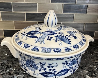 Pode incluir: Uma caçarola redonda de cerâmica com tampa, apresentando uma base branca e um padrão floral azul. A tampa tem um puxador em forma de ponta, e a base inclui duas pegas laterais. O design consiste em intrincados motivos botânicos em azul.