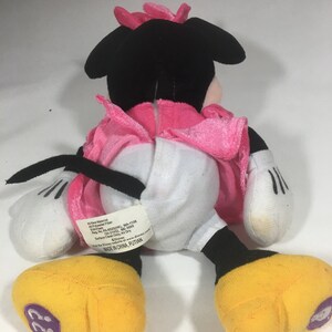May include: A black and white plush Minnie Mouse toy wearing a pink dress. The toy has a pink bow on its head and yellow shoes. The toy's tag reads "All New Material: Polyester Fiber,  Reg. No. PA-6520(CN), MA-1108,  Reg. No. PA-11422, MC-3605,  Surface Clean Only, Air Dry,  Disney, Visit the Disney website: www.disney.com, Made in China, PUTIAN"