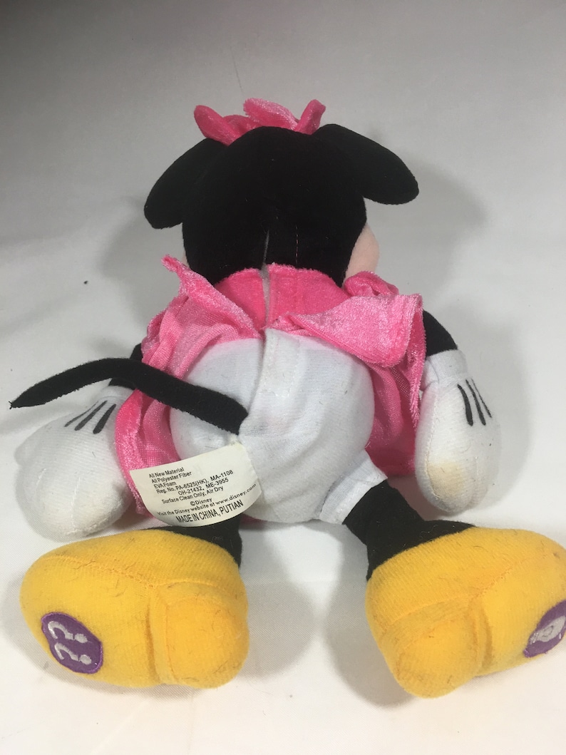 May include: A black and white plush Minnie Mouse toy wearing a pink dress. The toy has a pink bow on its head and yellow shoes. The toy's tag reads "All New Material: Polyester Fiber,  Reg. No. PA-6520(CN), MA-1108,  Reg. No. PA-11422, MC-3605,  Surface Clean Only, Air Dry,  Disney, Visit the Disney website: www.disney.com, Made in China, PUTIAN"