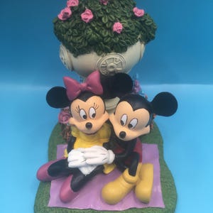 Puede incluir: Una figurita de cer&aacute;mica de Mickey Mouse y Minnie Mouse sentados en una manta morada con una base verde. Est&aacute;n sentados debajo de una maceta blanca con hojas verdes y flores rosas.