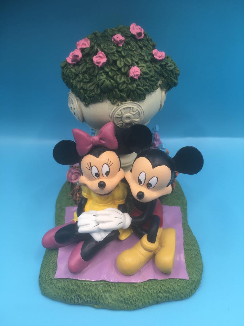 Puede incluir: Una figurita de cer&aacute;mica de Mickey Mouse y Minnie Mouse sentados en una manta morada con una base verde. Est&aacute;n sentados debajo de una maceta blanca con hojas verdes y flores rosas.