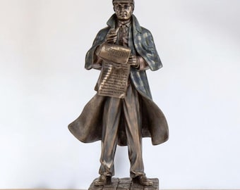 May include: A bronze-colored statue of a man in a hat and coat, holding a newspaper with the headline "MURDER." The statue is standing on a stone base, and the coat has a blue and green plaid pattern.