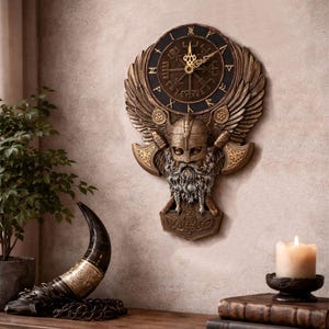 May include: A bronze-colored wall clock with a Viking theme. The clock features a round face with Roman numerals, a winged design, and a Viking helmet and axe motif. The clock is mounted on a beige wall.