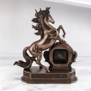 May include: Bronze-colored horse clock with a rearing horse design. The clock features a detailed sculpture of a horse with flowing mane and tail, leaning on a decorative clock face. The base has an ornate, scrolled design.