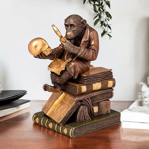 May include: A bronze sculpture of a chimpanzee, wearing a jacket, examining a golden skull with a magnifying glass. The chimp sits on a stack of books with gold detailing. The sculpture has a vintage, antique appearance.