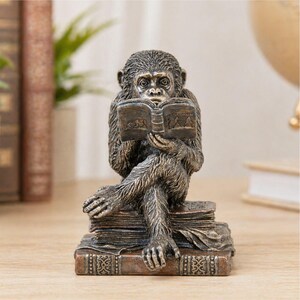 May include: A bronze-colored monkey figurine reading a book, seated atop a stack of books. The monkey has detailed fur and is engrossed in the book. The base of the figurine is a stack of books with decorative detailing.