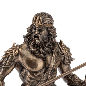 May include: Bronze-colored statue of a muscular man with a long beard, wearing a crown and armor. The statue has a stern expression and is holding a spear. The figure is a representation of a mythological god.