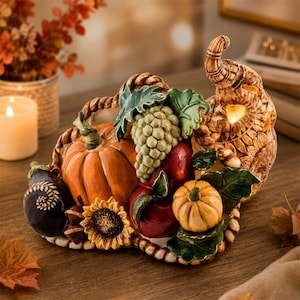 May include: A ceramic cornucopia centerpiece featuring a woven texture, filled with a sculpted orange pumpkin, a cluster of green grapes, a red apple, a small yellow pumpkin, a sunflower, and a dark purple eggplant, all accented with green leaves.