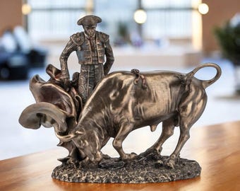May include: Bronze-toned sculpture depicting a bullfight scene. A matador stands near a charging bull, with a cape draped between them. The sculpture is detailed, with the figures and base showing texture and form. The background is blurred.