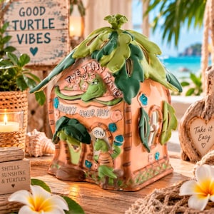 May include: A ceramic hut-shaped decorative piece with a peach-coloured base, green palm leaf roof, and sculpted turtle figures. The front features the text "THE TIPSY TURTLE" and "HAPPY HOUR HUT" with an "OPEN" sign above a small turtle figure.