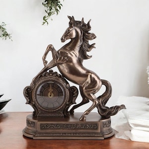 May include: Bronze-colored decorative clock featuring a rearing horse. The clock has an ornate, detailed design with a round clock face. The horse statue is positioned above the clock, creating a striking and elegant display. The clock sits on a rectangular base.