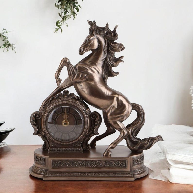 May include: Bronze-colored decorative clock featuring a rearing horse. The clock has an ornate, detailed design with a round clock face. The horse statue is positioned above the clock, creating a striking and elegant display. The clock sits on a rectangular base.
