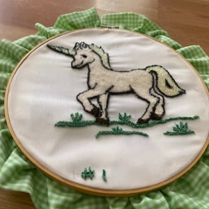 Può includere: Un unicorno bianco ricamato su un tessuto bianco teso su un cerchio di legno per ricamo. Il cerchio è circondato da un tessuto a quadri vichy verde e bianco.