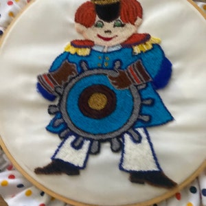 Può includere: Arte di ricamo a cerchio con un personaggio dei cartoni animati che indossa un'uniforme blu e oro, un cappello blu e tiene un volante blu. Il personaggio ha i capelli rossi e un viso sorridente. Il cerchio è decorato con un tessuto bianco con pois rossi, gialli e blu.