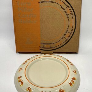 Op de afbeelding: Een ronde, gebroken witte keramische kaarsenhouder voor stompkaarsen met een herhalend patroon van maissnoepjes langs de rand en een oranje cirkelvormige rand. Het item staat voor de originele bruine kartonnen doos met de tekst Candy Corn Pillar Candle Holder.