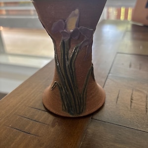 May include: A terracotta-colored ceramic vase with a flared rim and base. The exterior features a raised, sculpted relief of a purple iris flower with green stems and leaves extending from the base toward the top of the vessel's body.