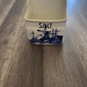 May include: A white ceramic salt container with a blue and white landscape design featuring a windmill and the word "SALT". The container has a rectangular shape with a curved top and a hole for hanging.