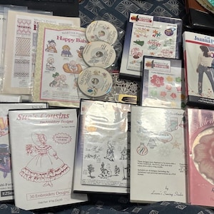 May include: A collection of embroidery design software packages and pattern booklets featuring various themes including Christmas trees, floral motifs, and character illustrations. The items are presented in plastic cases and paper sleeves with printed titles and graphics.