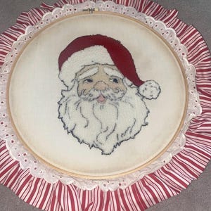 Può includere: Un'opera d'arte a ricamo con Babbo Natale. L'immagine presenta un volto dettagliato di Babbo Natale con cappello rosso e barba bianca. Il ricamo è incorniciato in un telaio di legno, circondato da un bordo di pizzo bianco e tessuto a righe rosse e bianche.
