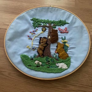 Può includere: Opera d'arte ricamata in un cerchio di legno, con sfondo azzurro. Il disegno raffigura un albero con tre orsi marroni, un alveare e oggetti volanti colorati. Gli orsi sono su un'area erbosa verde.