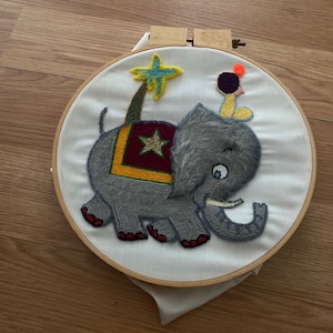 Può includere: Arte di ricamo a cerchio che presenta un elefante grigio con una sella a motivi rosso, giallo e verde, una stella gialla e un piccolo uccello con la testa nera e il becco arancione.