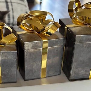 May include: Three grey metal gift boxes with gold ribbon and accents. The boxes are arranged in a row, each of a different size. The gold ribbon is wrapped around the boxes like a present.