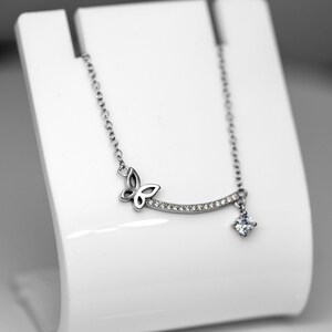 May include: A silver-toned necklace featuring a butterfly design and a dangling square-cut gemstone. The necklace has a delicate chain and a curved bar adorned with small, clear gemstones. The butterfly is on the left side of the curved bar.