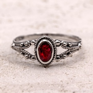 May include: A silver ring featuring an oval, deep red gemstone. The ring has an intricate design with beaded and braided details. The gemstone is set in a silver bezel. The ring is displayed against a neutral background.