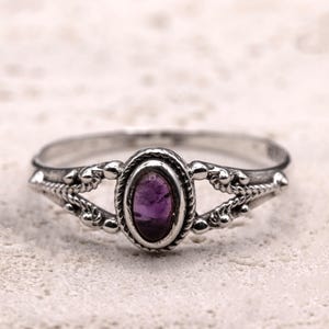 Puede incluir: Un anillo de plata con una piedra preciosa ovalada de color púrpura oscuro. El anillo tiene un diseño detallado y ornamentado con un borde trenzado alrededor de la piedra y detalles decorativos en la banda. Es una joya.