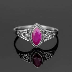 以下が含まれることがあります： 鮮やかなピンク色のマーキス型宝石をあしらったシルバーリング。宝石の周りとバンドに沿って、装飾的なフィリグリーデザインが施されています。リングは反射する黒い表面に表示され、その複雑なディテールが際立っています。