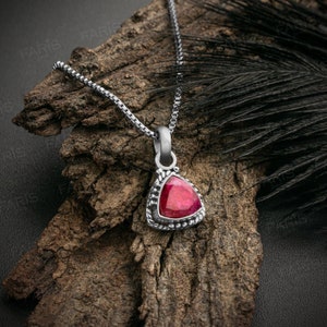 May include: A silver chain necklace with a triangular pendant featuring a red gemstone. The pendant has a detailed silver border.