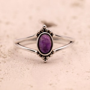 Op de afbeelding: Een zilveren ring met een ovale, paarse edelsteen. De ring heeft een gesplitst bandontwerp en decoratieve kralenaccenten rond de edelsteen. De ring is geplaatst tegen een neutrale achtergrond.