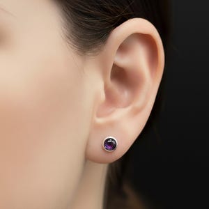 Peut inclure: Gros plan sur une oreille avec une petite boucle d'oreille à clou en améthyste ronde. La boucle d'oreille a un sertissage argenté et une pierre précieuse violet foncé. Le fond est de couleur sombre.