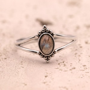 May include: A silver ring with an oval gemstone in shades of brown, grey, and blue. The ring has a split band and decorative bead detailing around the gemstone. The ring is shown on a light-coloured surface.