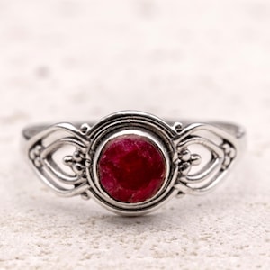 May include: A silver ring featuring a central, round, deep red gemstone. The ring has an intricate, openwork design with curved lines and small beaded details. The gemstone is set within a circular bezel.