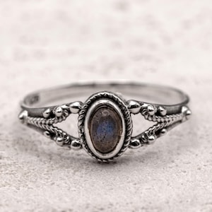 Può includere: Un anello d'argento con una gemma ovale e iridescente. L'anello presenta un design ornato con dettagli perlati e intrecciati attorno alla gemma e sulla fascia. La gemma ha sfumature blu e grigie.