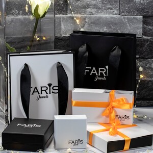 May include: Four white gift boxes with black ribbon and the text "Faris Jewels" printed on them. The boxes are arranged in a pyramid shape, with the largest box at the bottom and the smallest box at the top. The boxes are sitting on a gray brick surface.