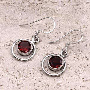 May include: A pair of silver earrings with a circular design. Each earring features a deep red gemstone set within a silver circle. The earrings have a hook closure.