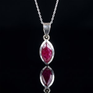 May include: A silver pendant necklace featuring a marquise-cut ruby gemstone. The ruby is a deep red color and set in a silver bezel. The pendant hangs from a delicate silver chain. The background is black, creating a contrast that highlights the jewelry.