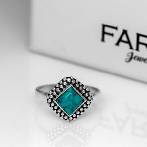 May include: A silver ring with a turquoise gemstone set in a square-shaped bezel. The bezel is surrounded by small silver beads.