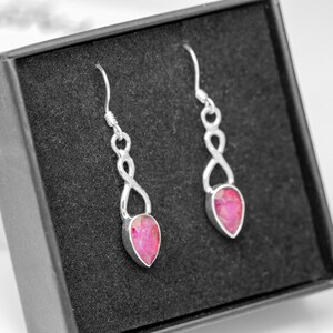 May include: A pair of silver-colored drop earrings with a teardrop-shaped, pink gemstone. The earrings have an infinity symbol design and are displayed in a black jewelry box.