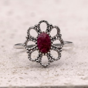 Puede incluir: Un anillo de plata con un engaste en forma de flor. El engaste tiene un diseño de cuentas en los bordes y sostiene una gema ovalada de color rojo intenso. El anillo está sobre una superficie texturizada de color blanquecino.