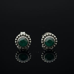 May include: A pair of silver stud earrings with a dark green, round gemstone center. The earrings have a decorative, beaded border. The earrings are displayed on a reflective black surface.
