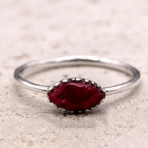 Puede incluir: Un anillo de plata con una piedra preciosa roja oscura con corte marquesa. La piedra preciosa está engastada en un bisel decorativo. La banda del anillo es delgada y pulida, creando un diseño minimalista. El anillo se muestra sobre una superficie texturizada de tono neutro.