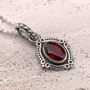 May include: A silver pendant necklace featuring a deep red oval gemstone set in an ornate, detailed frame. The pendant has a decorative bail and hangs from a delicate chain. The design is vintage-inspired.