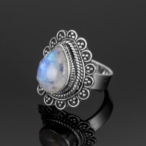 May include: A silver ring with a teardrop-shaped moonstone set in a detailed silver bezel. The ring has a decorative band with a rope-like design and a scalloped edge.