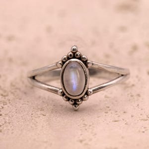 Op de afbeelding: Een zilveren ring met een ovale, iriserende edelsteen. De ring heeft een gesplitst bandontwerp en is versierd met kleine, parelachtige details rond de edelsteen setting. De ring wordt getoond op een getextureerd, lichtgekleurd oppervlak.