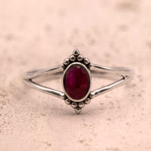 Pode incluir: Um anel de prata com uma pedra preciosa ovalada de cor vermelha escura. O anel tem um design de haste dividida e detalhes decorativos de contas ao redor da pedra preciosa. O anel está sobre um fundo neutro e texturizado.